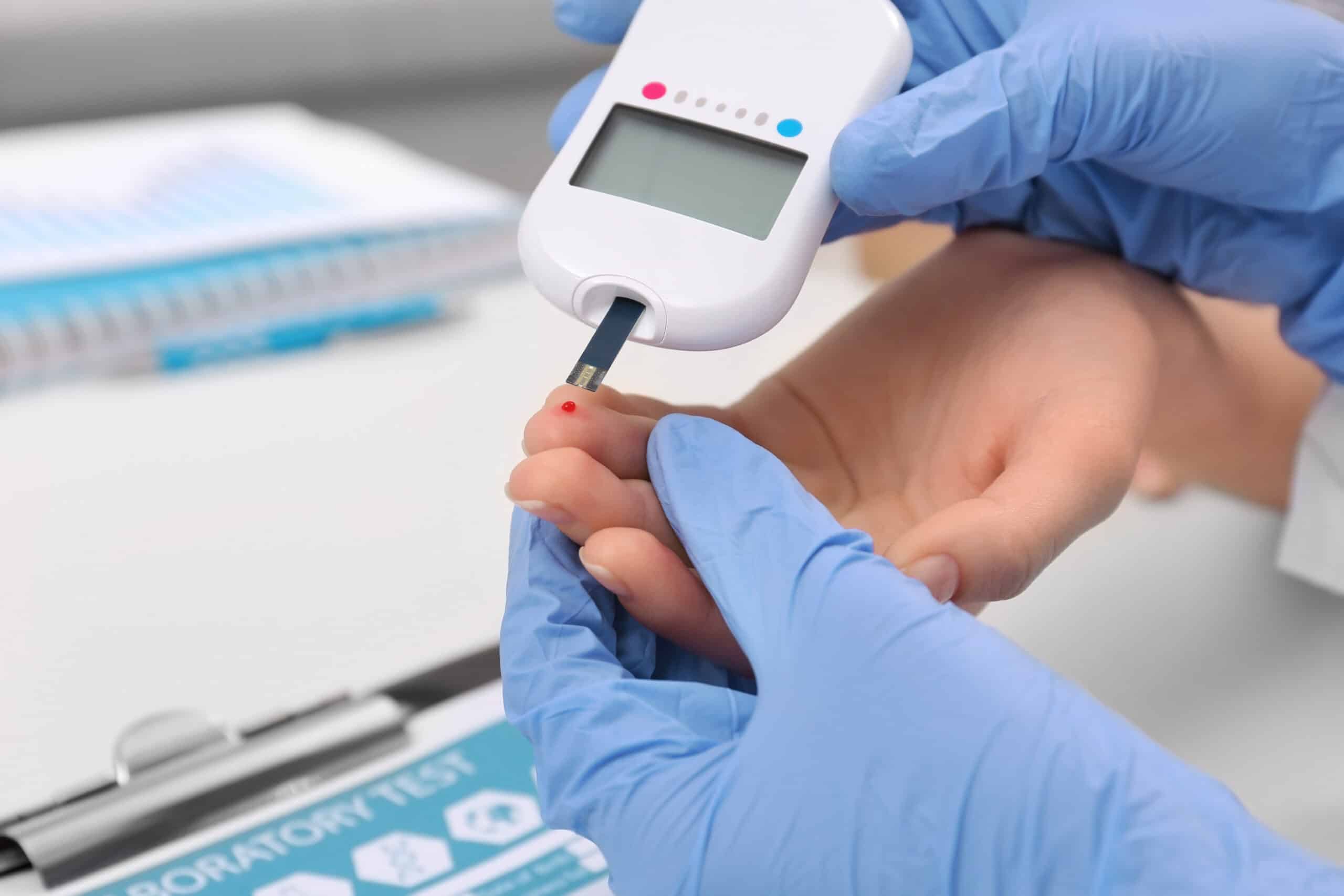 A blue gloved provider’s hand testing blood sugar from a patient’s finger.